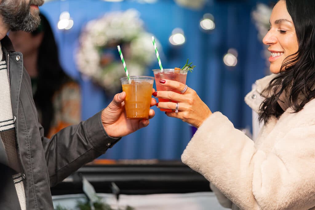 Two people clinking glasses with cocktails at Winterfest wearing winter coats, man and woman.